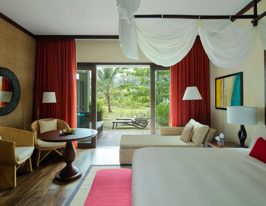 Elegant and spacious resort room with modern decor, large bed with white canopy, and a view of the tropical gardens through floor-to-ceiling windows.