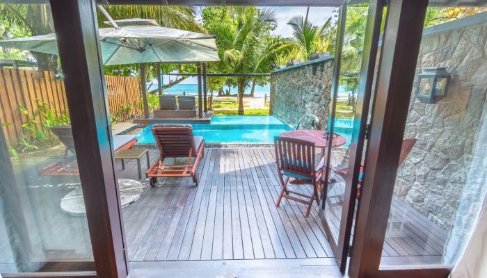 View from a Seychelles resort suite showing a cozy wooden deck with sun loungers, a pool, and lush palm trees leading to a tranquil beachfront.