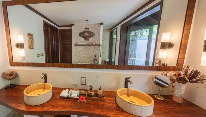 A modern bathroom with twin stone basins on a wooden countertop, toiletries, and a large mirror reflecting a bright, airy room with a tropical view through the glass door.