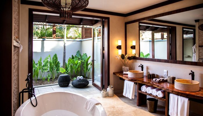 A Seychelles resort bathroom with a freestanding bathtub, wooden vanity with twin stone basins, wall-mounted mirrors, and a private garden view with lush tropical foliage.