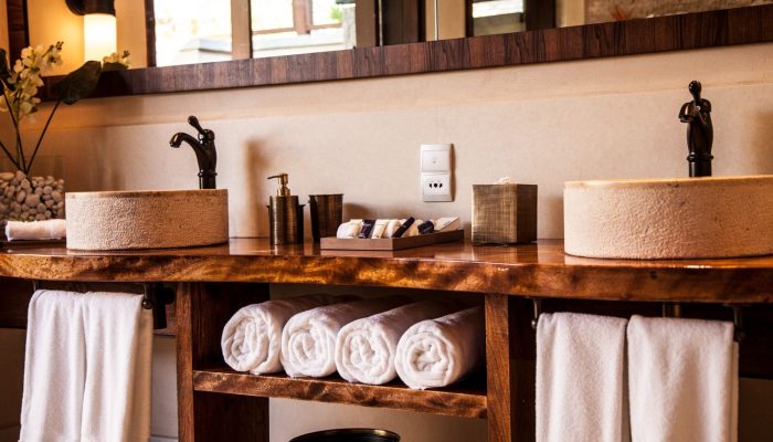 A hotel bathroom counter with two circular stone sinks, a wooden shelf with white towels, and a small mirror.