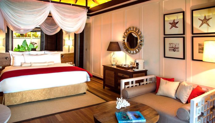 Elegant Seychelles hotel room featuring a draped four-poster bed, contemporary furnishings, marine-themed wall art, and warm lighting.