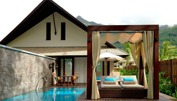 A private poolside cabana at a luxury resort in Seychelles, with a draped four-poster daybed, two loungers with turquoise cushions, and a siting area in the background.