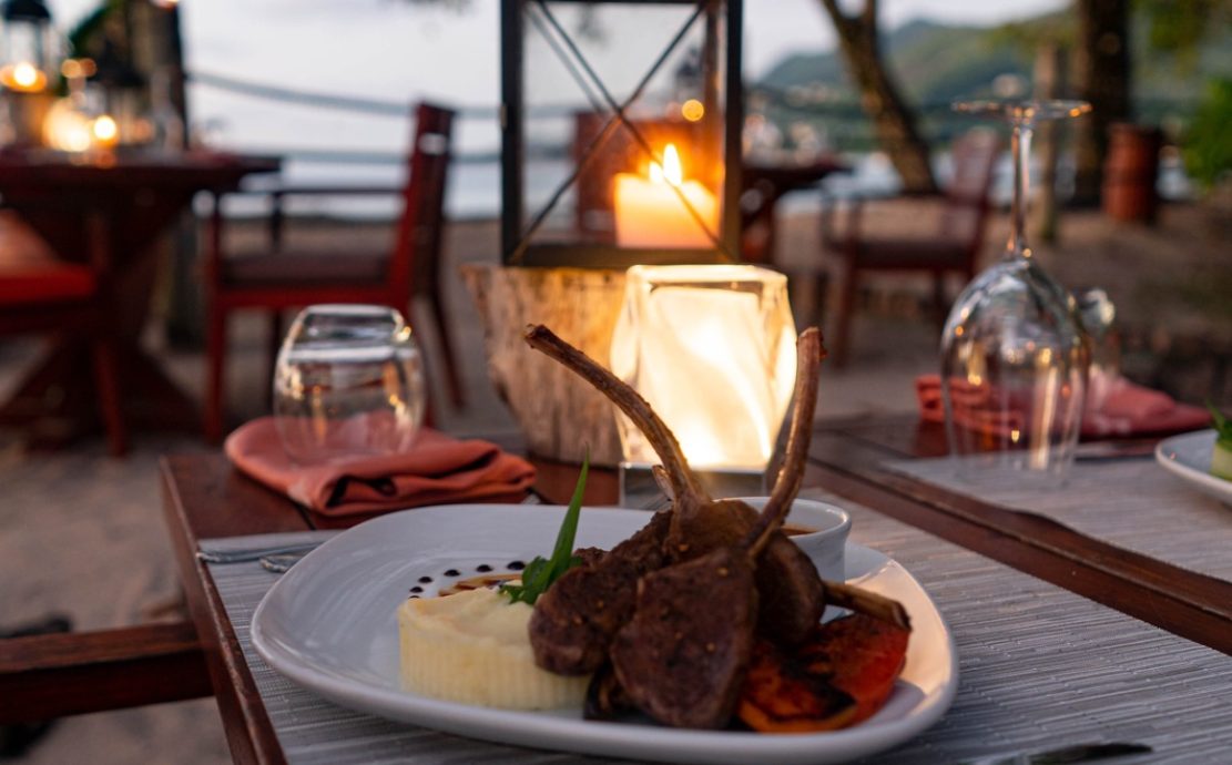 Romantic beachside restaurant in Seychelles with a plate of lamb chops and a glowing lantern on a rustic table at dusk.