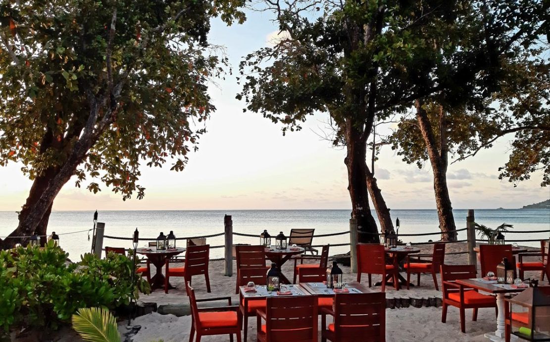 Dawn at Eden, a beachfront restaurant on Beau Vallon Beach, with inviting tables amidst tropical trees overlooking the calm Seychelles sea.
