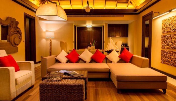 Warmly lit Seychelles hotel suite with plush sofas adorned with red cushions, intricate wood carvings, and a high beamed ceiling.