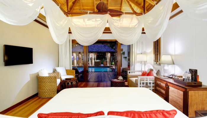 A luxurious Seychelles hotel suite with a king-size bed, sheer canopy draping, tropical wooden ceiling and furniture, and sliding glass doors leading to a private pool.