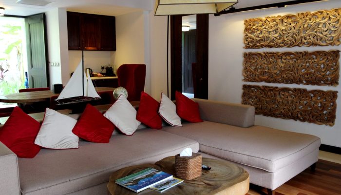 Elegant lounge area of a Seychelles hotel suite, featuring a plush sectional sofa with red and white pillows, a wooden coffee table, and a sailboat model.
