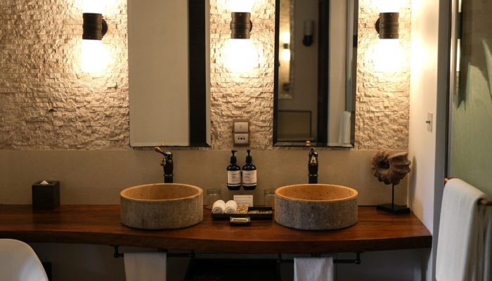 Warmly lit bathroom in a resort suite featuring dual stone basins, rectangular mirrors, mounted wall lights, and a textured stone wall.