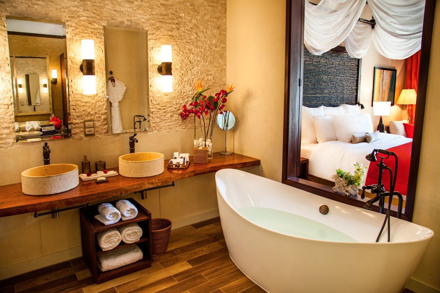 A suite's luxurious bathroom with dual stone basins, a standalone bathtub, and a reflection of the bedroom with white draping and red accents in Seychelles.