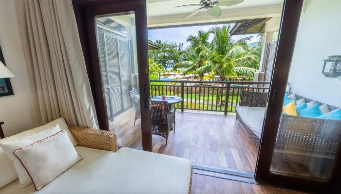 Resort room in Seychelles with a plush sofa, a glass door opening to a balcony with a view of palm trees, and tropical décor.