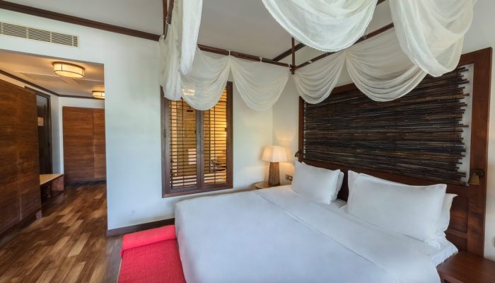 Elegant bedroom inside a hotel in Seychelles with a large bed, billowing white curtains, warm lighting, a dark wood headboard, and a red accent rug.