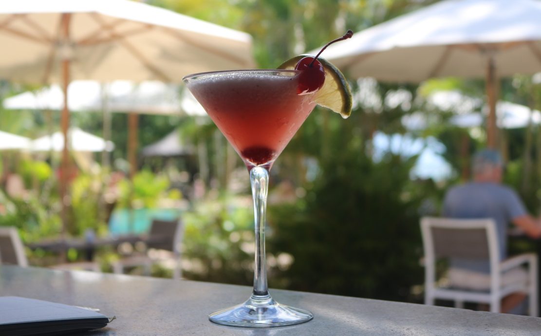 Chilled cocktail with a cherry garnish on an outdoor bar counter with poolside umbrellas in the background.