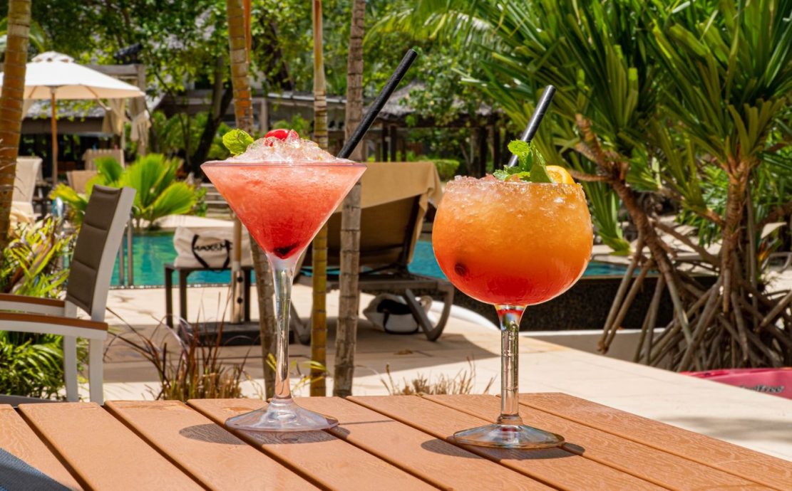 Two frosted glasses of slushy tropical drinks on a poolside table in a Seychelles resort, with sun loungers and umbrellas in the background.