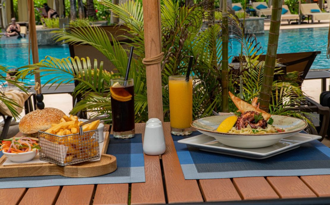 Poolside lunch featuring a cheeseburger with fries, a seafood dish, and refreshing beverages at a Seychelles resort.