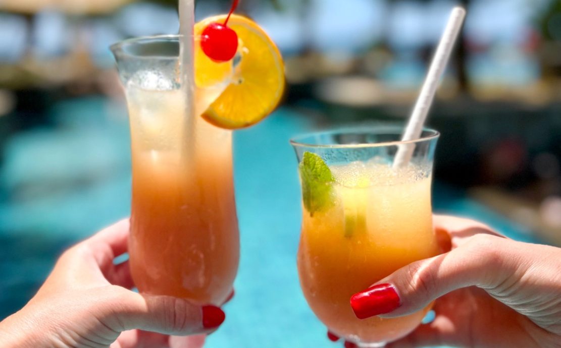 Two hands holding tropical cocktails by a resort pool in the Seychelles.