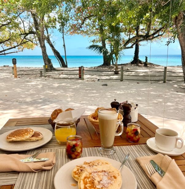 A breakfast spread for two with pancakes, pastries, fruit salad, coffee, and a latte set on a beachfront table on the Beau Vallon Beach.
