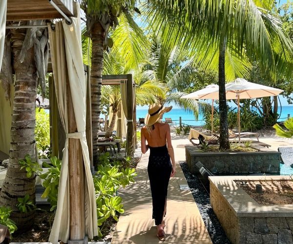 Woman strolling towards the beachside cabanas lined with palm trees on a sunny day in the Seychelles.
