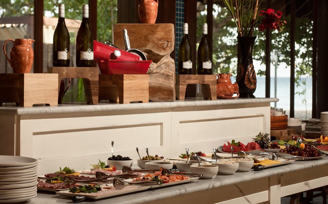Luxurious buffet spread with a variety of cheeses, cold cuts, and fresh fruits, complemented by bottles of Prosecco, set against a backdrop of Beau Vallon Beach.