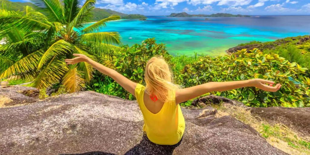 A person sitting on a rock with arms outstretched, overlooking the stunning turquoise waters of Seychelles Islands.