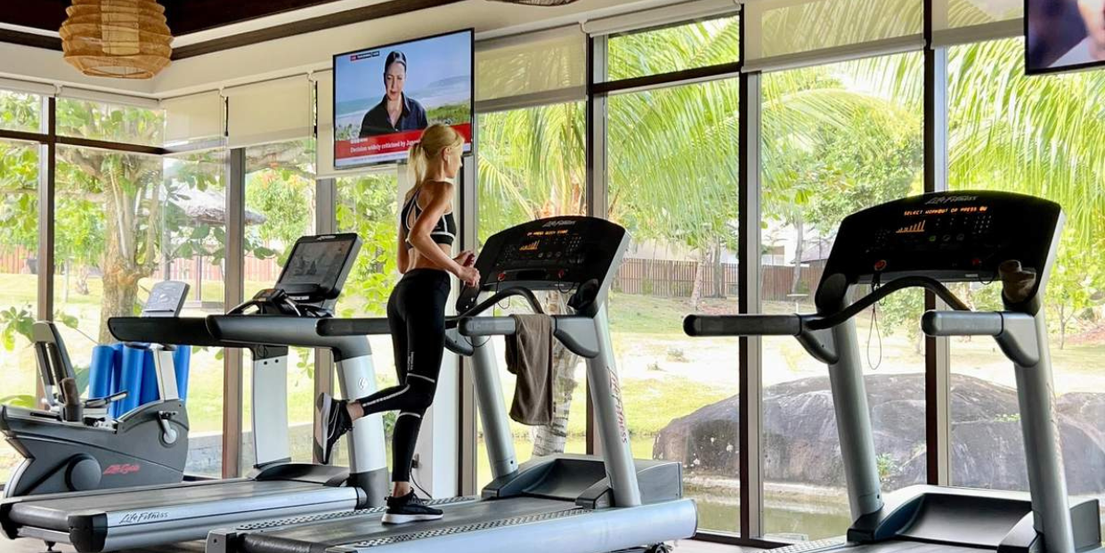 Fitness enthusiast using a treadmill in a well-equipped Seychelles gym at a luxury resort, with a view of the tropical landscape outside.
