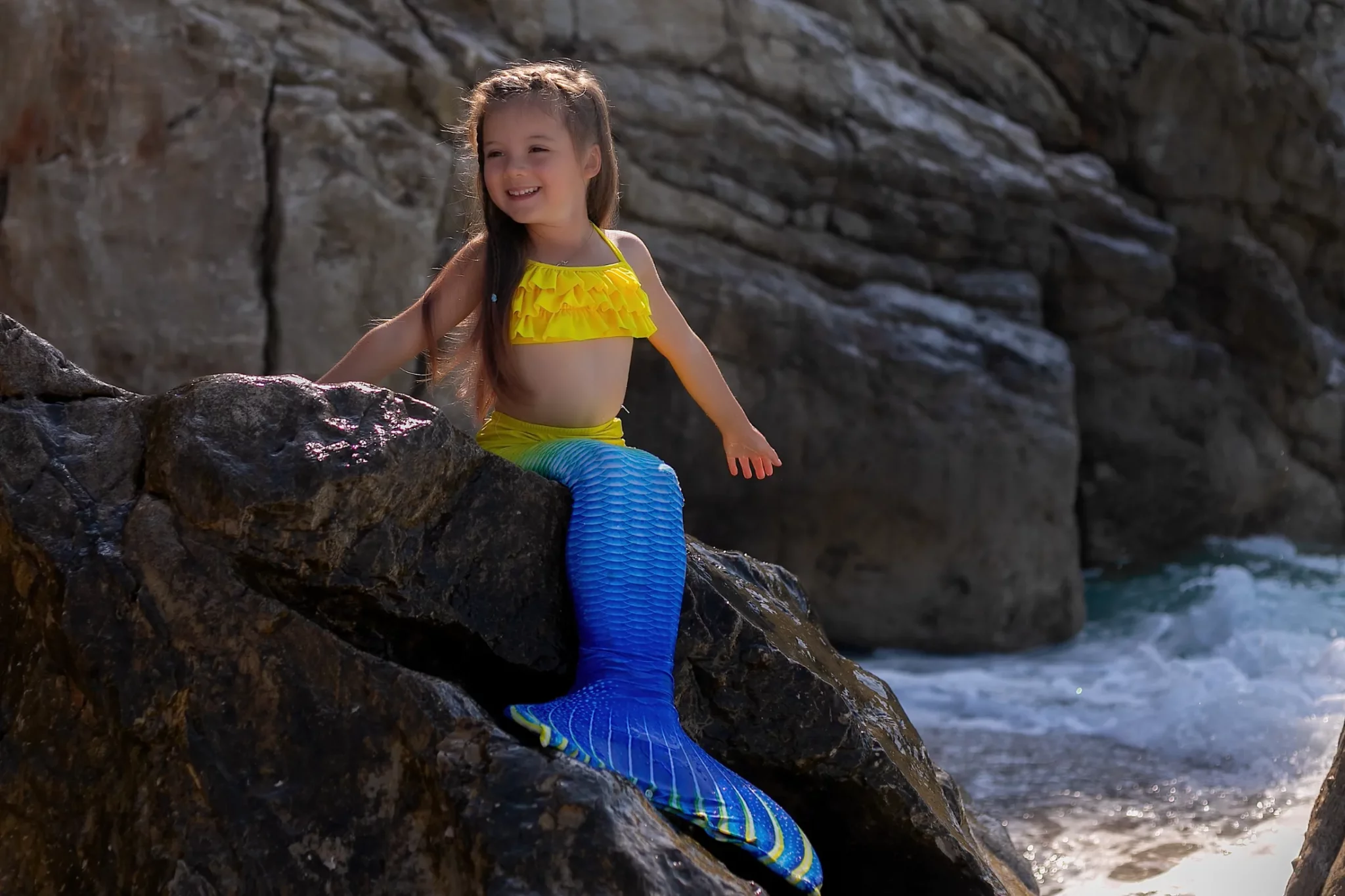 A smiling young girl in a mermaid costume sitting on a rock by the sea.