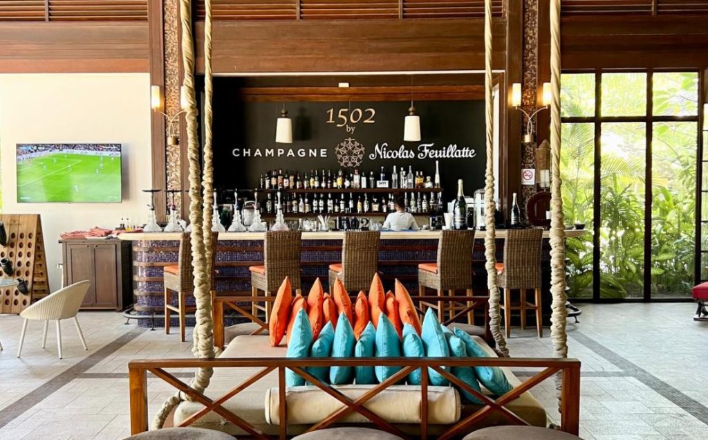 Inside the 1502 Bar in Seychelles with a stylish interior, featuring a well-stocked bar, wicker bar stools, and vibrant cushions.