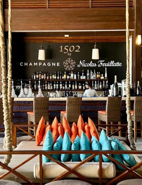 Inside the 1502 Bar in Seychelles with a stylish interior, featuring a well-stocked bar, wicker bar stools, and vibrant cushions.