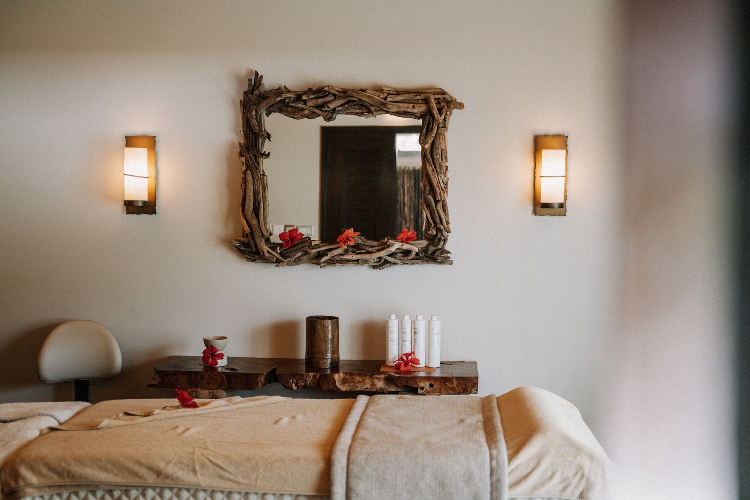A spa treatment room with a rustic wooden mirror, wall sconces, massage table, and products on a shelf.
