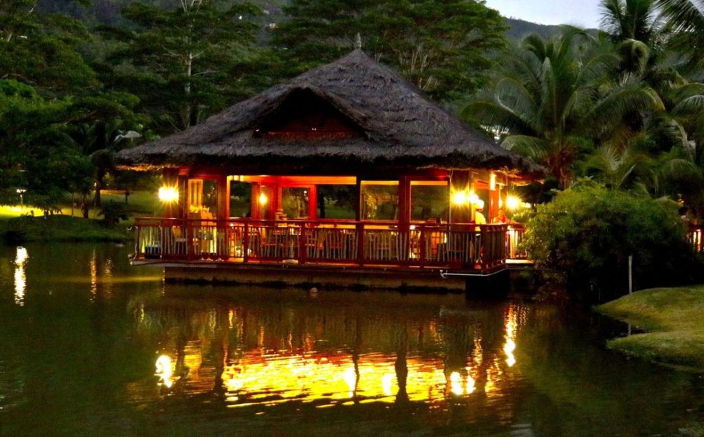 Thatched-roof gazebo illuminated at night, reflecting on the calm waters of a tropical resort in the Seychelles.