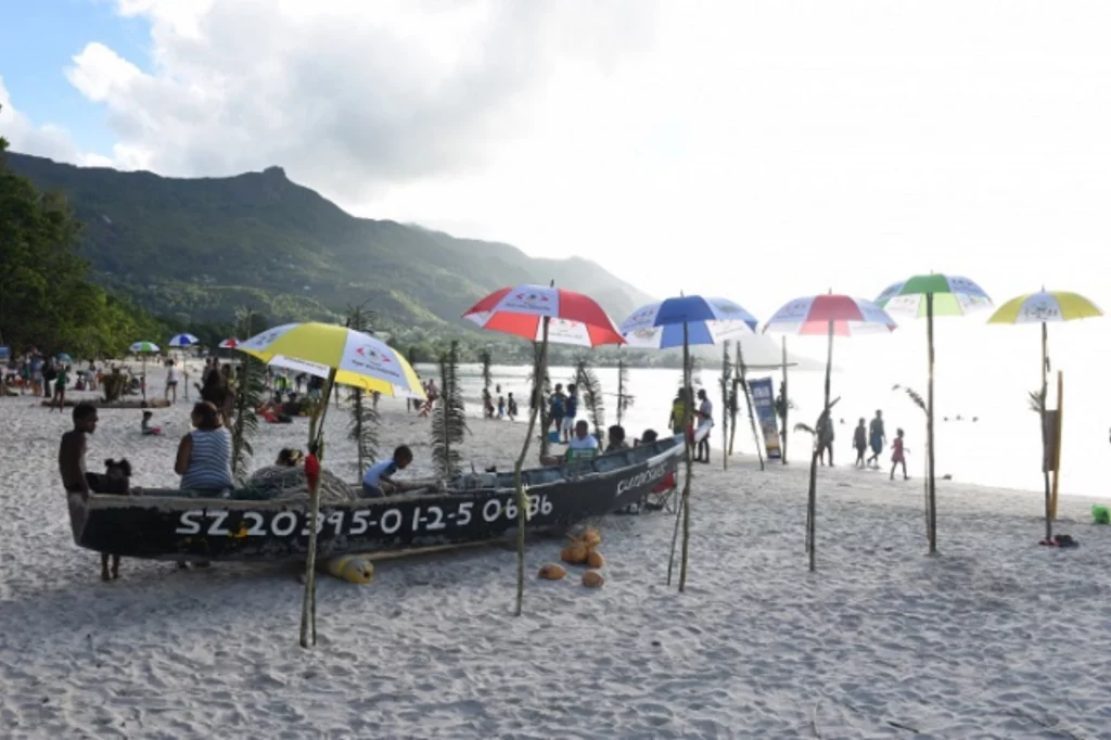 Crowded beach with colorful umbrellas and a traditional boat on the sand, set against a backdrop of lush mountains in the Seychelles.
