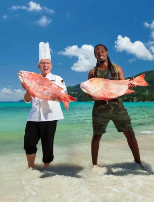 A chef and a fisherman proudly holding large, fresh fish on the shore, showcasing the local catch against the backdrop of a beautiful Seychelles beach.