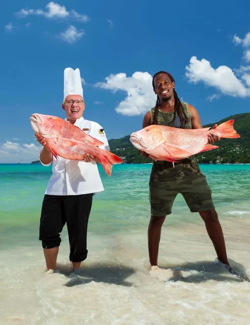 A chef and a fisherman proudly holding large, fresh fish on the shore, showcasing the local catch against the backdrop of a beautiful Seychelles beach.