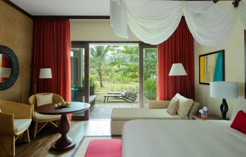 Elegant and spacious resort room with modern decor, large bed with white canopy, and a view of the tropical gardens through floor-to-ceiling windows.